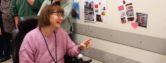 Ung kvinde i kørestol smiler og peger på et billede på en opslagstavle